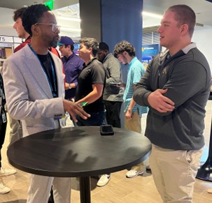 Two people stand talking at a high-top table during a networking event, with other attendees conversing in the background.