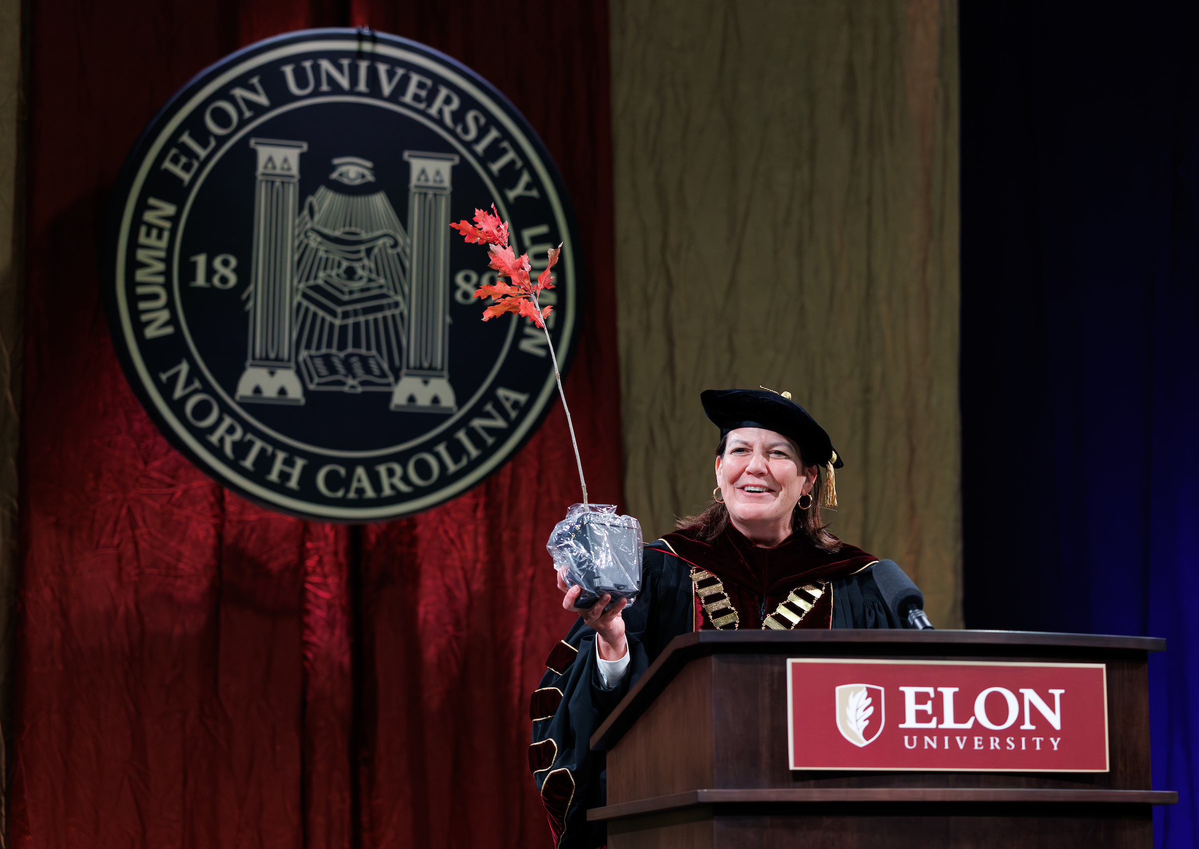 Connie Ledoux Book in academic regalia holding an oak sapling and speaking at a podium.