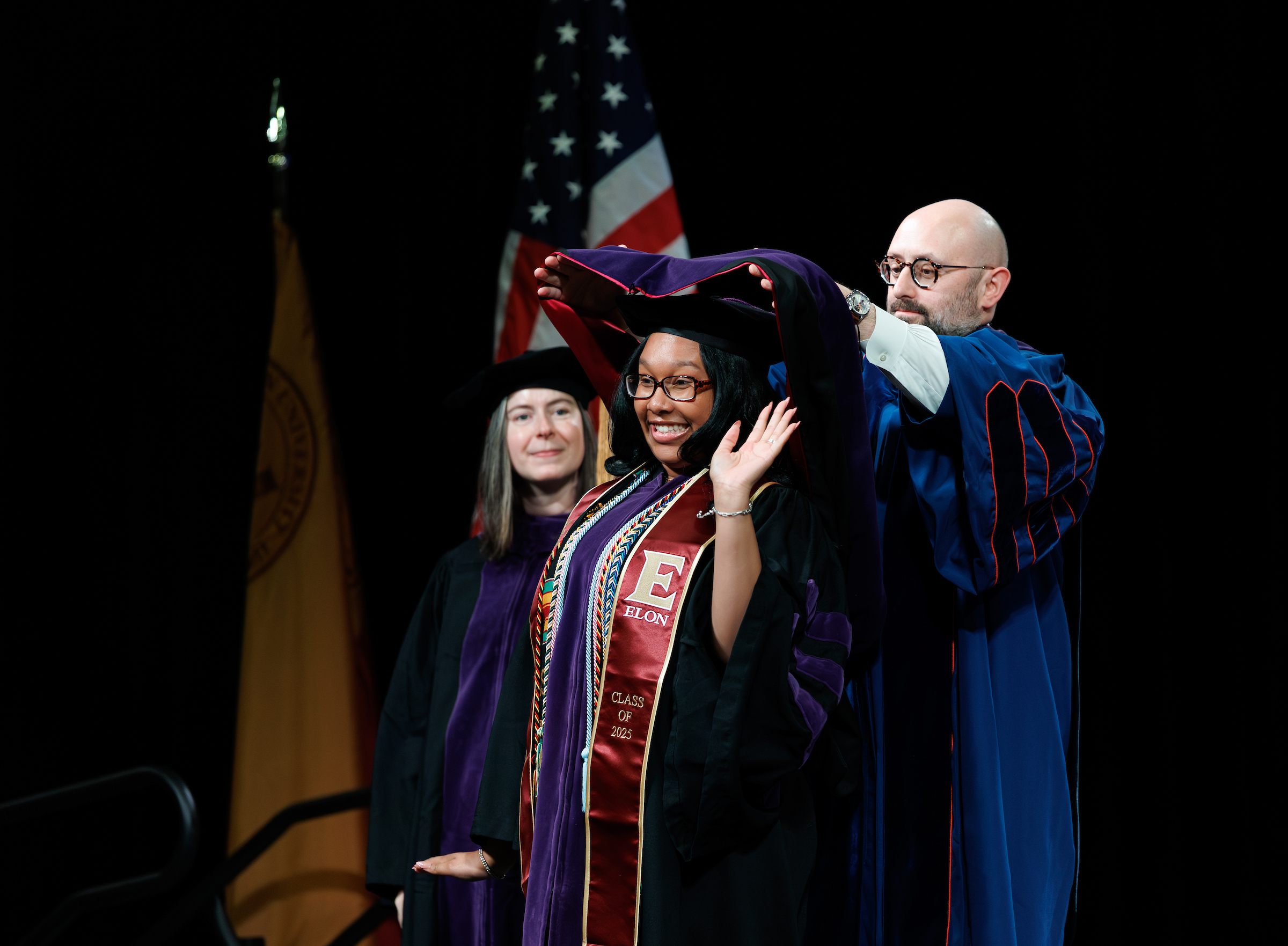 A woman being hooded by a dean smiles and waves at friends and family in the audience