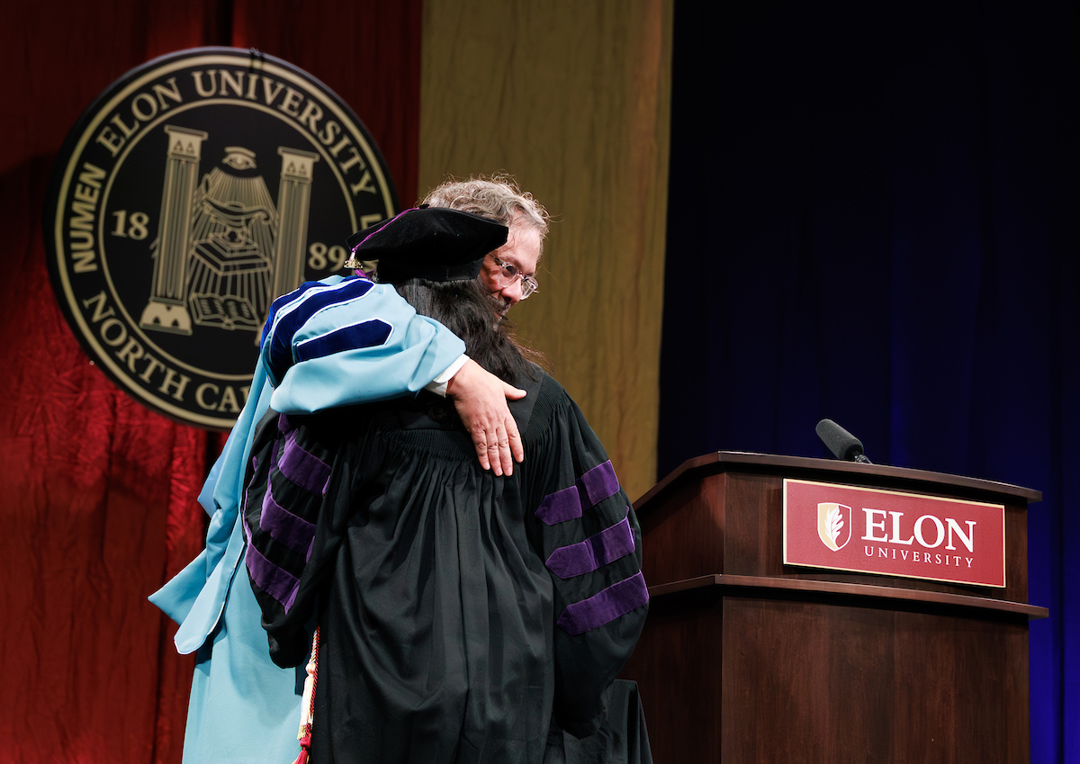 A professor and student in academic regalia hug by a podium that says Elon University.