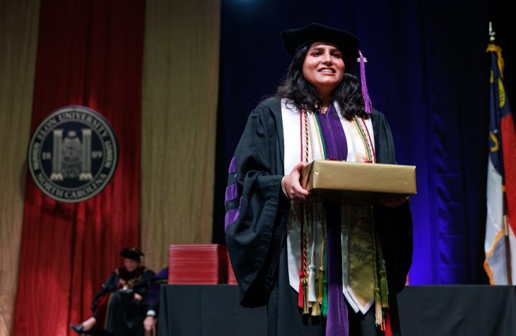 A woman in academic regalia carrying a gift-wrapped box across a Commencement stage