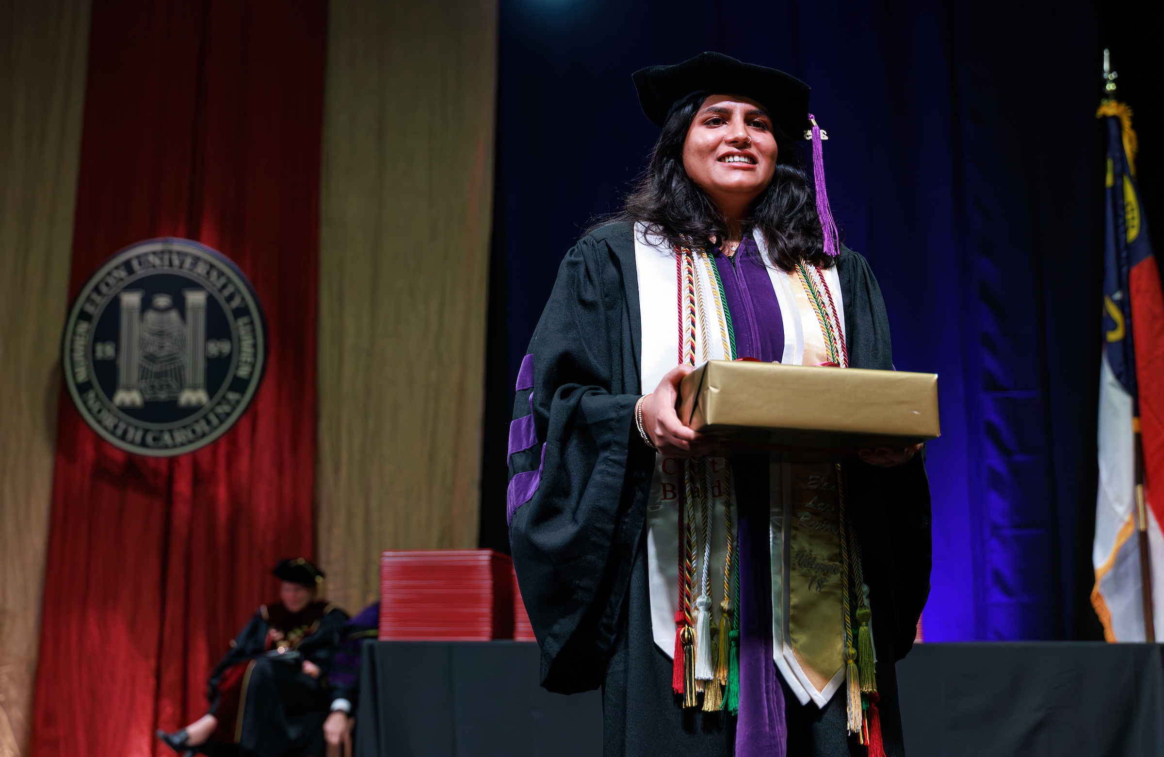 A woman in academic regalia carrying a gift-wrapped box across a Commencement stage