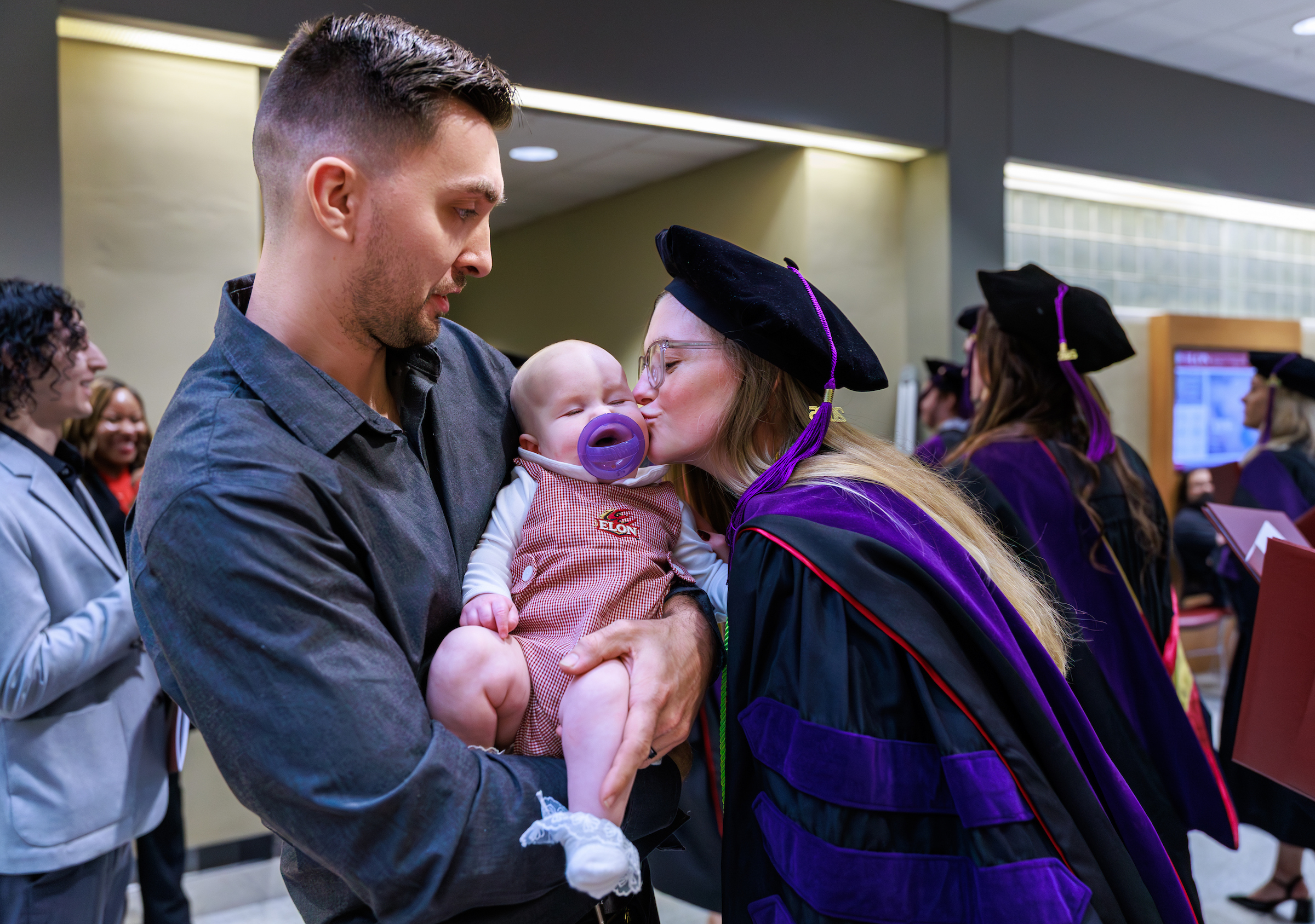 A woman in academic regalia kisses a sleeping baby held by her husband.