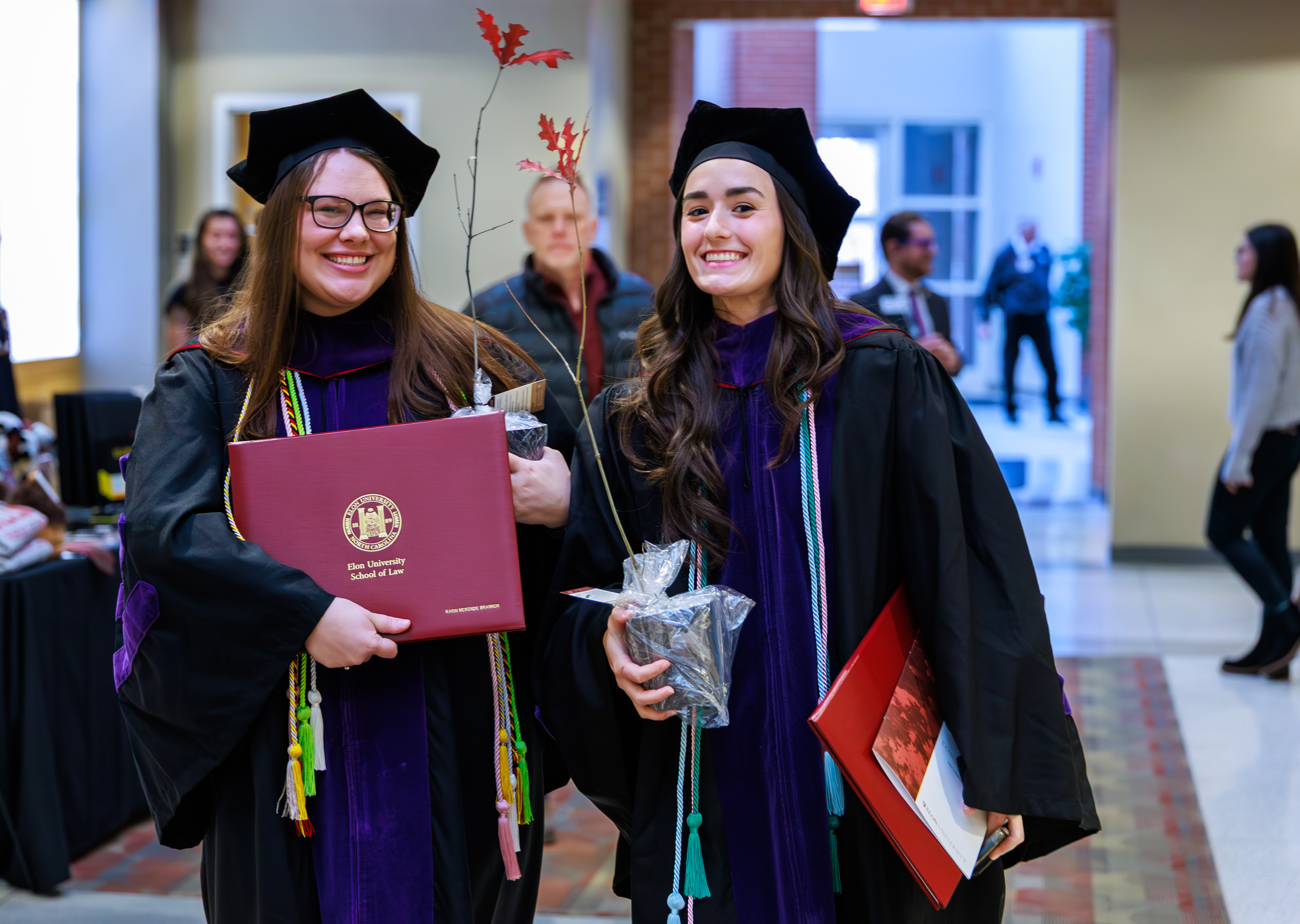 Two women students in academic regalia smiling, holding diplomas and saplings in the conocourse of Alumni Gym