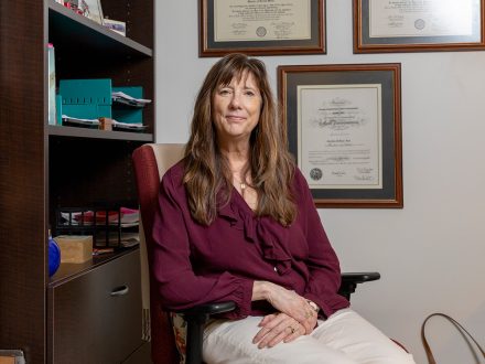 A faculty member sits in an office chair, smiling gently toward the camera, with shelves and framed diplomas visible behind her.