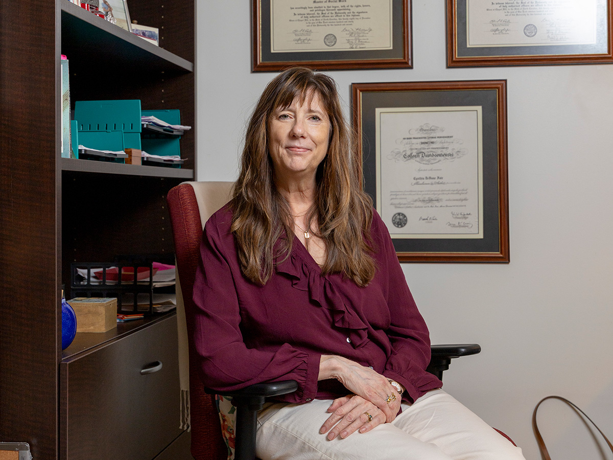 A faculty member sits in an office chair, smiling gently toward the camera, with shelves and framed diplomas visible behind her.