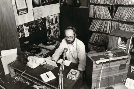 A black-and-white photograph shows a radio broadcaster seated at a desk surrounded by records and audio equipment.
