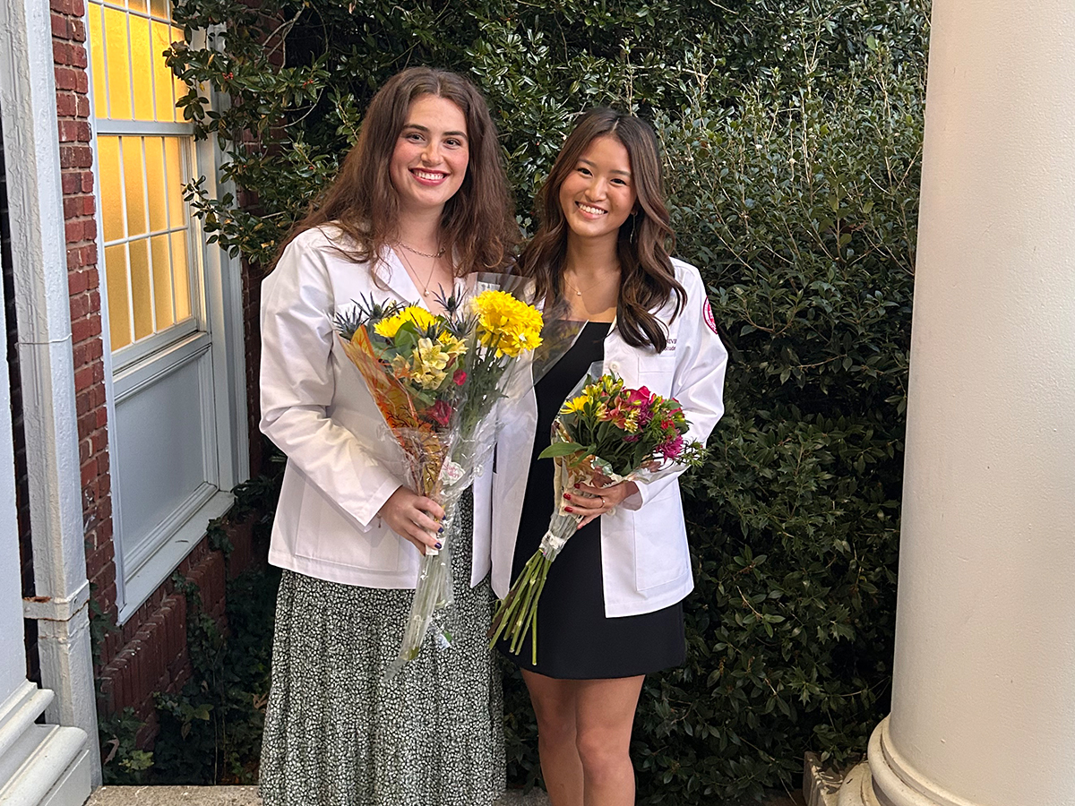Two people in white coats stand outside between a brick building and a column, each holding a bouquet of flowers. They smile as they pose together in front of leafy greenery.