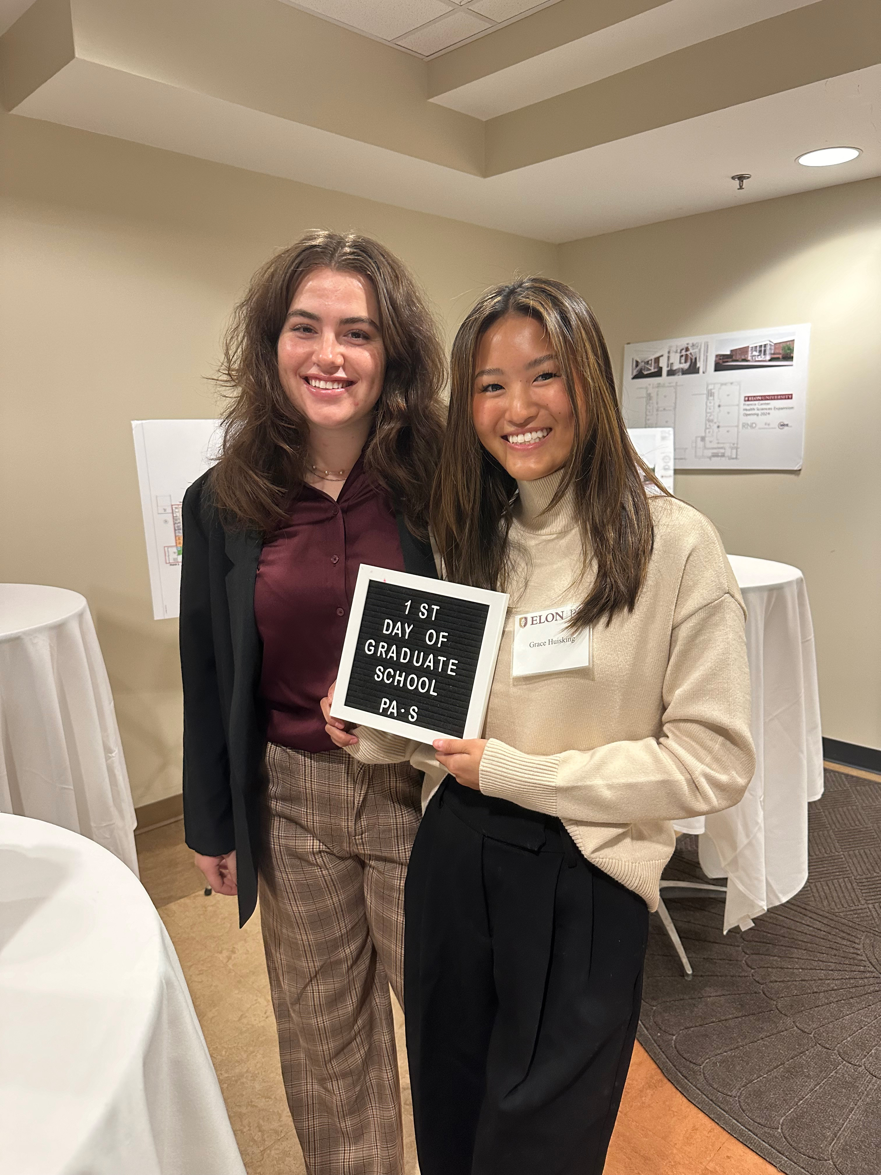 Two people stand indoors smiling, with one holding a small sign that reads “1st Day of Graduate School PA-S.” Round tables with white tablecloths and wall displays are visible in the background.
