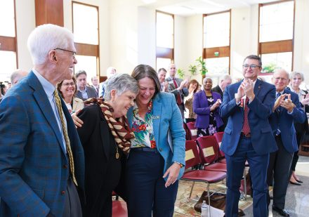 University leaders and guests applaud as two women share an emotional embrace during a campus celebration event.