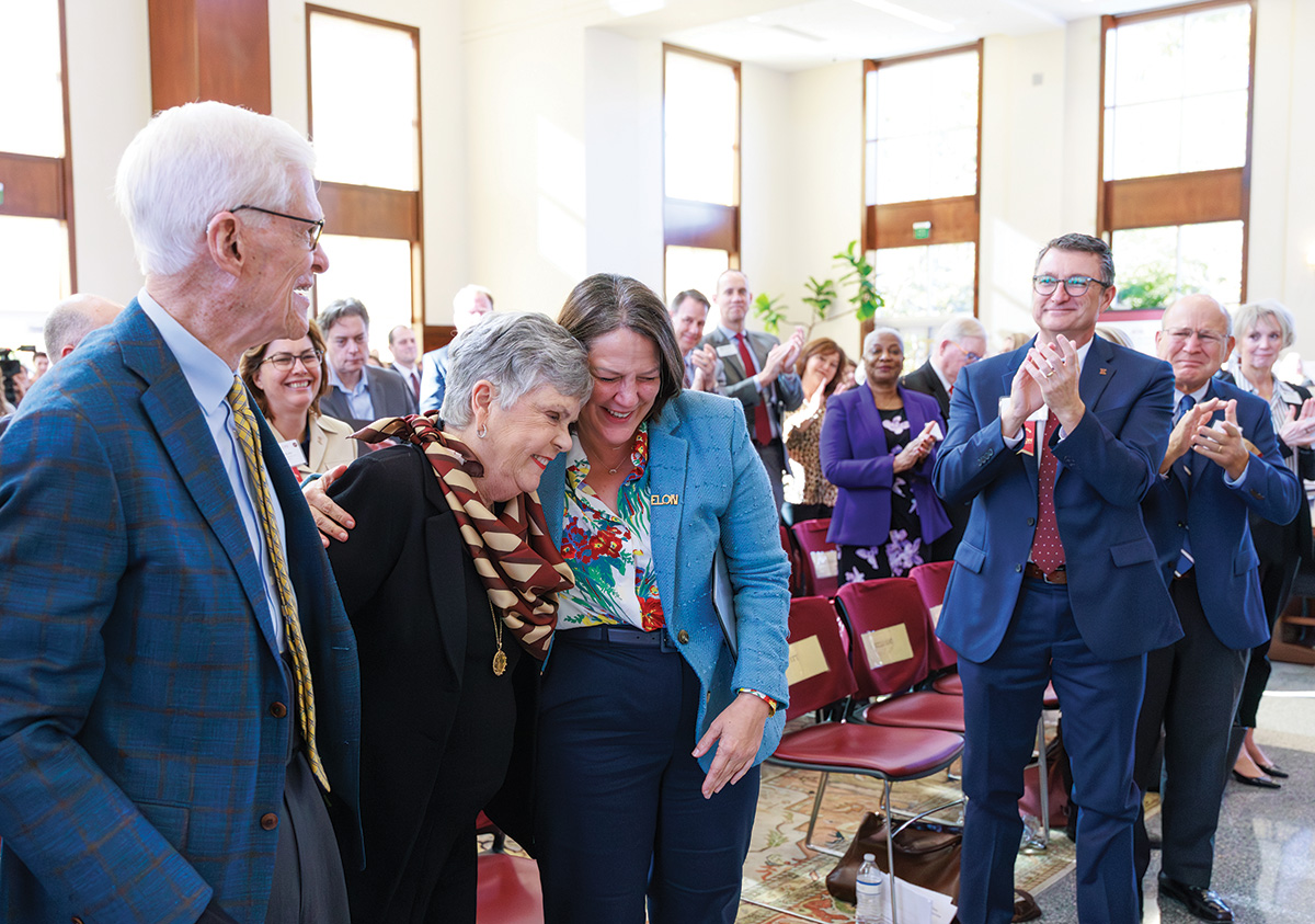 University leaders and guests applaud as two women share an emotional embrace during a campus celebration event.