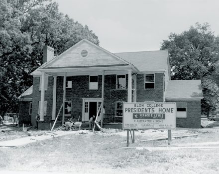 A black-and-white photograph shows the construction of the Elon College president’s home, with workers and scaffolding visible.
