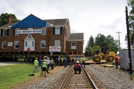 Workers oversee the relocation of a brick house marked “Elon University Recycles” as it is transported across railroad tracks.