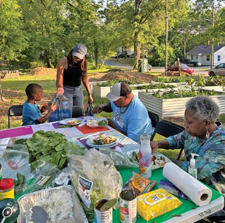 Adults and children sit together at an outdoor table preparing and eating food made with fresh vegetables.
