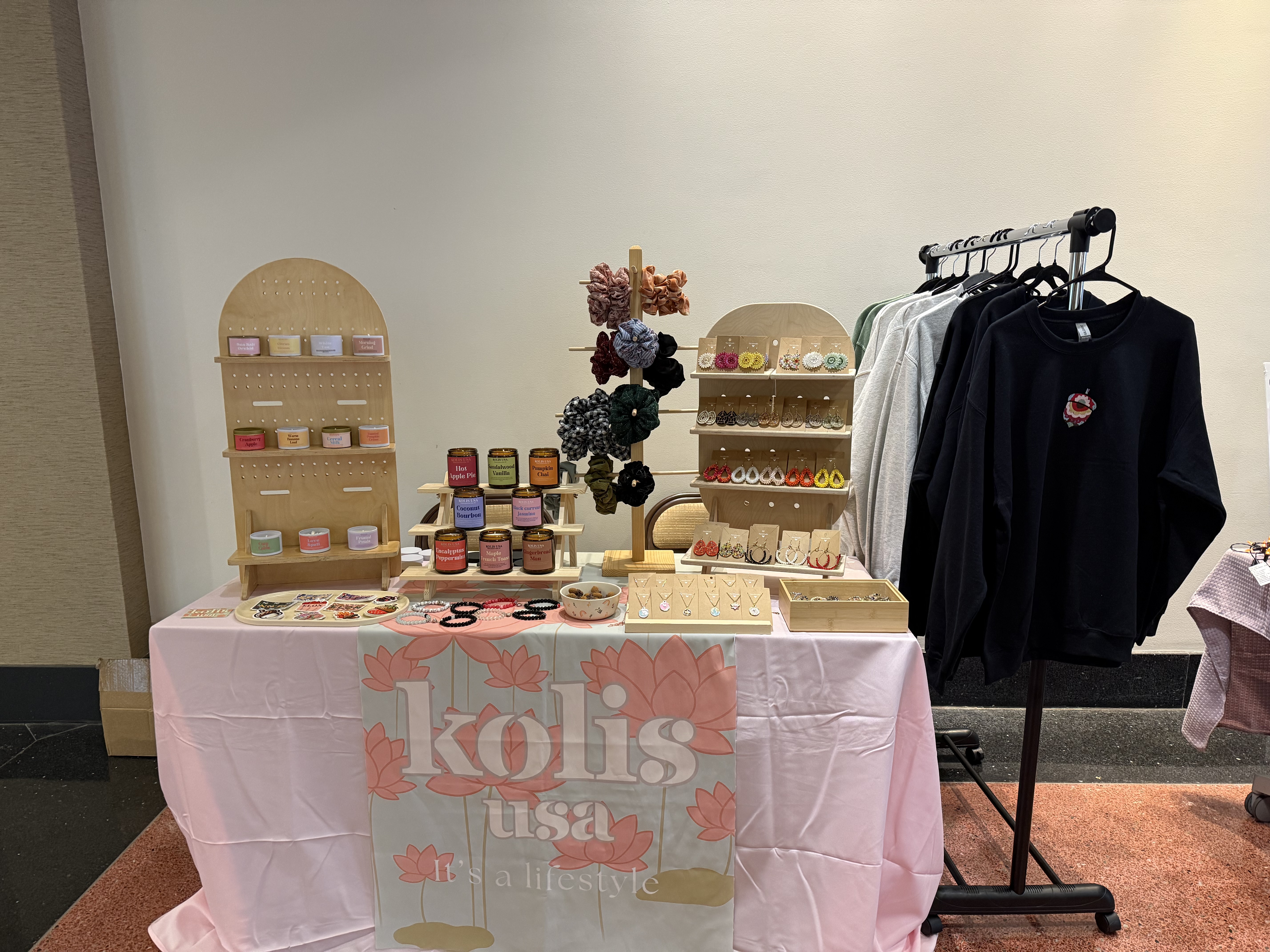 A vendor table displays colorful handmade items including candles, earrings, scrunchies, stickers, and necklaces, with sweatshirts hanging on a rack beside it and a banner reading “kolis usa.”