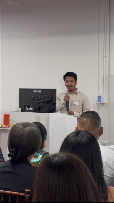 Man standing and holding microphone at a podium speaking to a group of attendees sitting in front of him.