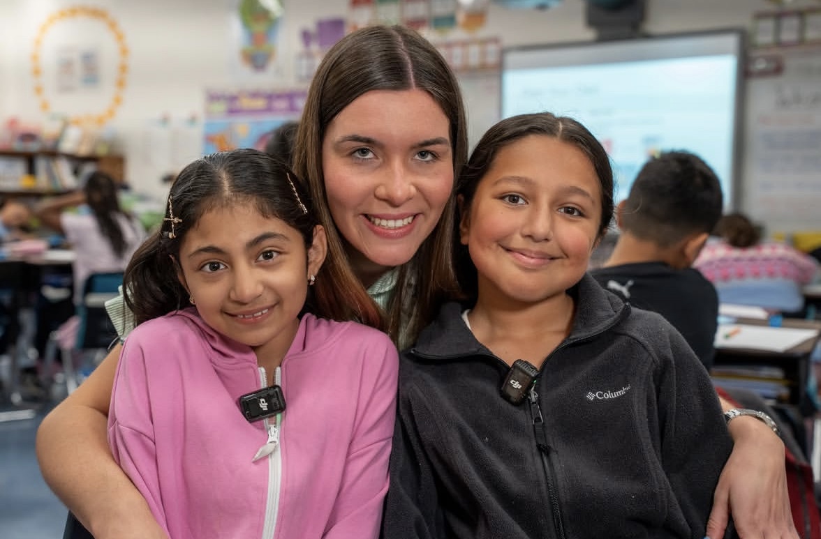 A teacher stands between two students in a classroom, smiling with her arms around them as they pose closely together.