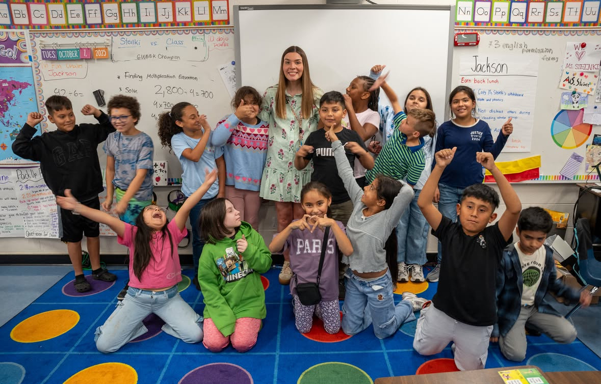 A teacher stands at the center of a classroom surrounded by excited students striking playful poses on a colorful rug.