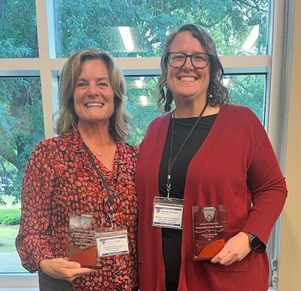 Two smiling women stand indoors in front of a large window with trees visible outside. Both wear conference name badges and hold glass awards. The woman on the left wears a red patterned blouse, and the woman on the right wears glasses, a black top, and a red cardigan.