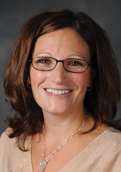 A female professor wearing a tan shirt and glasses smiles at the camera in a professional headshot.