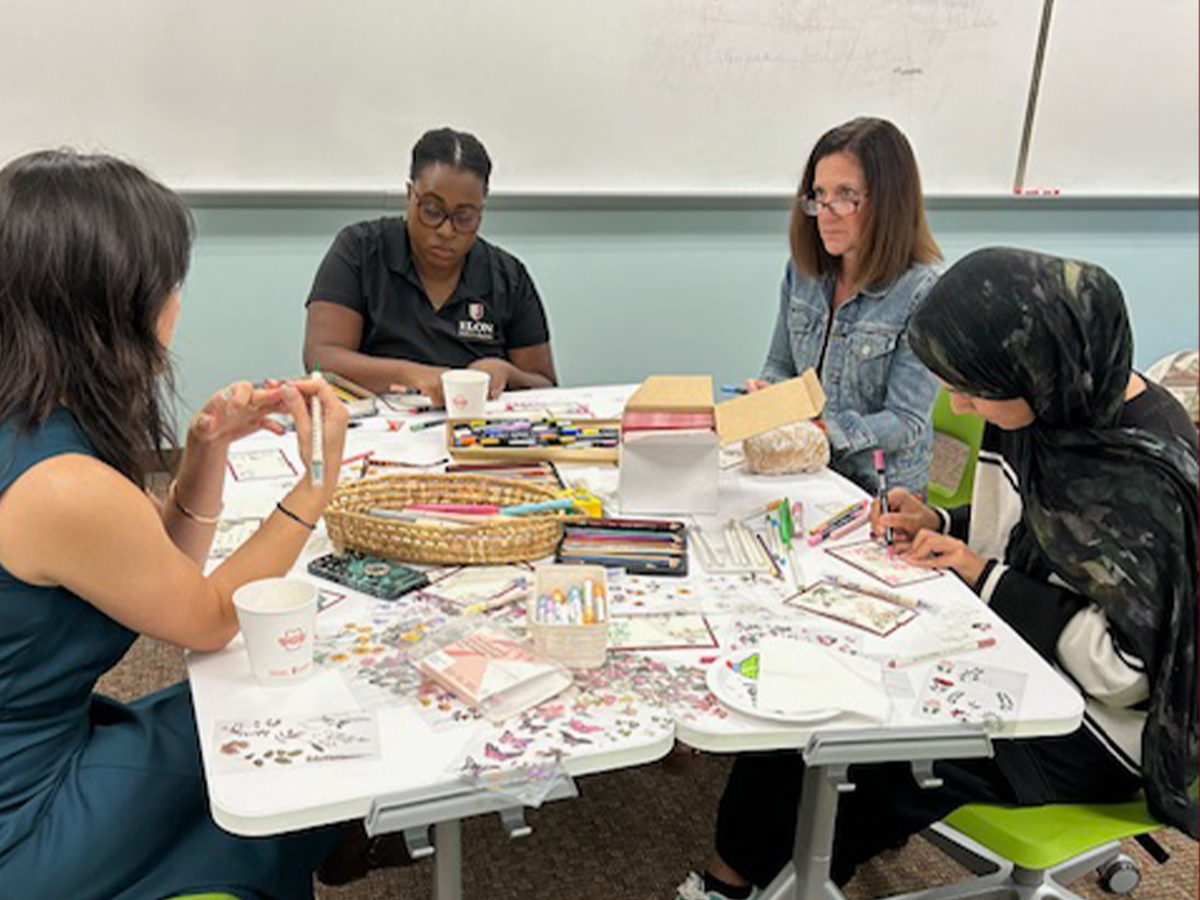 Four people sit around a table covered with art supplies, writing and decorating postcards together in a classroom setting.