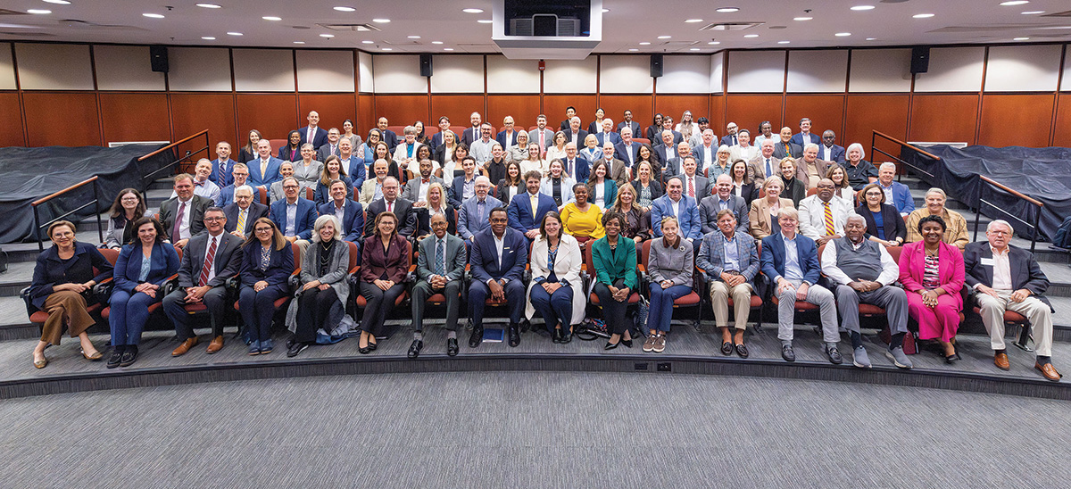 A large group of trustees and university leaders sit and stand together for a formal group photo inside a campus auditorium.