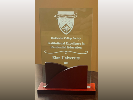 Glass award with a wooden base recognizing Elon University for ‘Institutional Excellence in Residential Education’ from the Residential College Society, dated 2025. The award features the RCS shield logo and decorative scrollwork