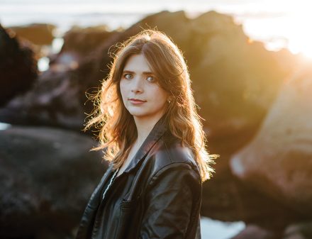 A portrait of a young woman wearing a leather jacket standing in front of a rocky coastline.