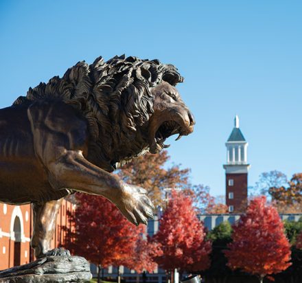 The Rex the Lion statue overlooks the Queens University campus in autumn, with colorful trees and the clock tower rising behind him.