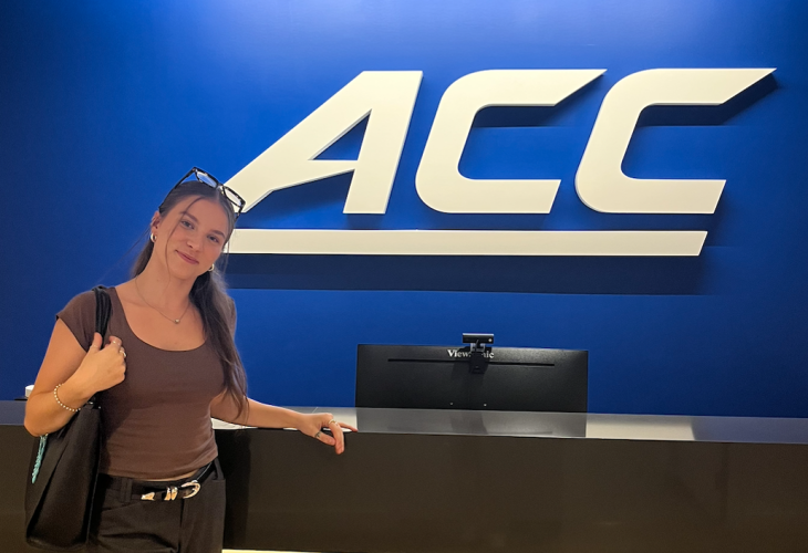 A person stands smiling in front of a bright blue wall with a large ACC logo, leaning lightly on a counter with a monitor behind it.
