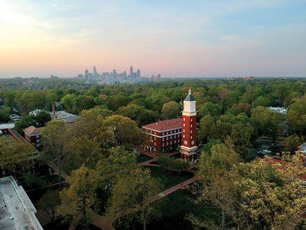 An aerial view of Queens University’s campus shows the clock tower rising above trees, with the Charlotte skyline in the distance.
