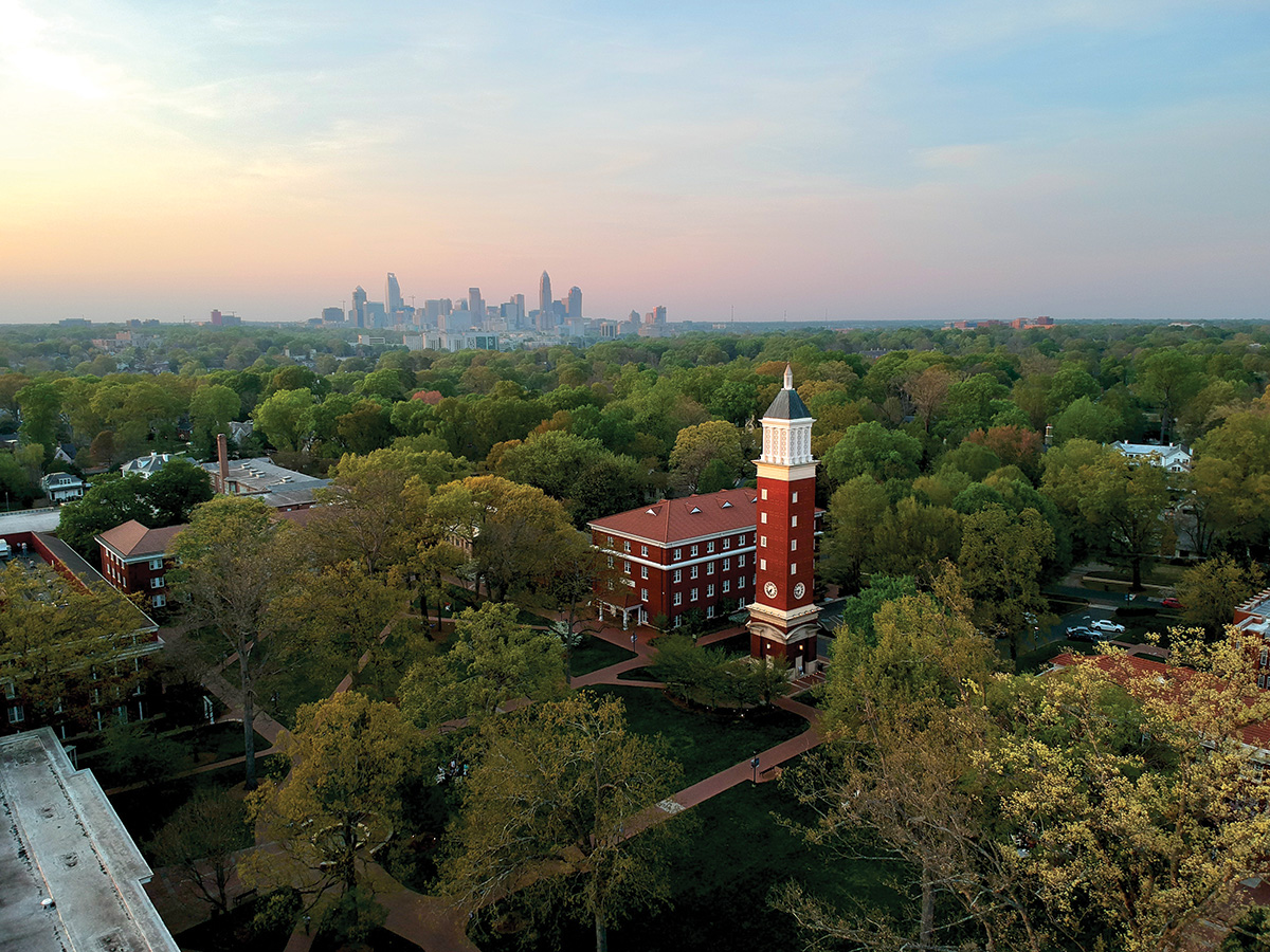 An aerial view of Queens University’s campus shows the clock tower rising above trees, with the Charlotte skyline in the distance.