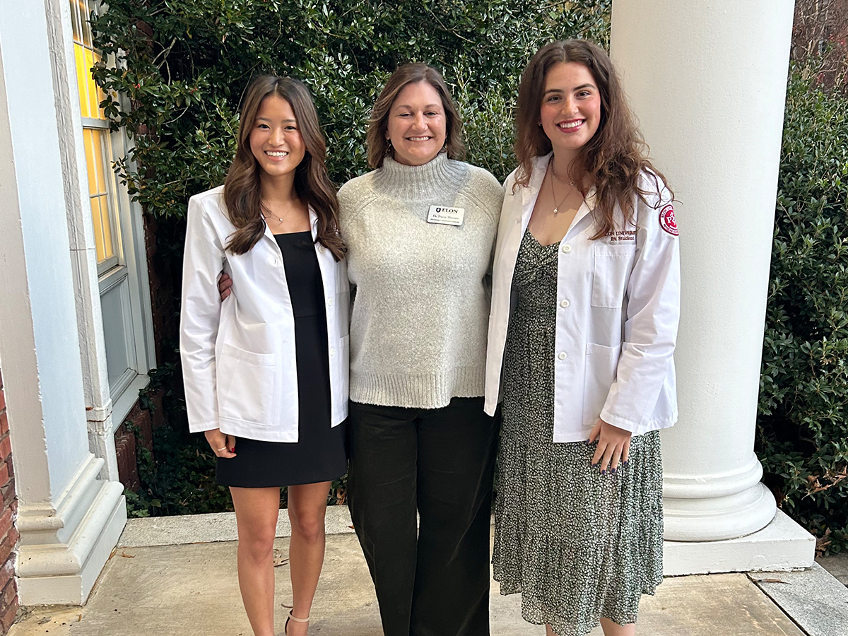 Three people stand outside in front of a column and greenery, with the two on the ends wearing white coats. They smile at the camera with their arms around each other.