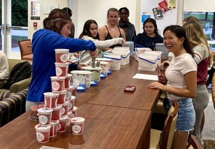 Students gather around a long table during a social event, smiling as one student scoops ice cream from large tubs. Stacks of red Italian ice cups and containers of sprinkles sit on the table, creating a build-your-own sundae station. The group appears relaxed and cheerful in a lounge area.