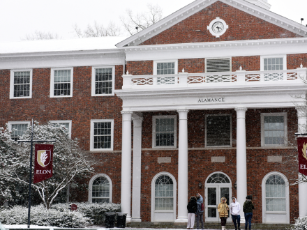 Snow falls in front of Alamance Building at Elon University, covering the roof, trees, and ground in a light layer of white. Several students bundled in winter clothing stand and talk near the building’s columned entrance as Elon banners hang on lamp posts nearby.