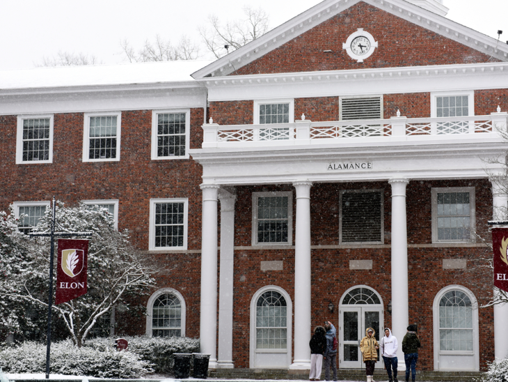 Snow falls in front of Alamance Building at Elon University, covering the roof, trees, and ground in a light layer of white. Several students bundled in winter clothing stand and talk near the building’s columned entrance as Elon banners hang on lamp posts nearby.