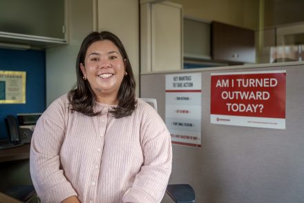 A person sits smiling in an office cubicle, wearing a light pink sweater, with posters on the wall behind them including one that reads “AM I TURNED OUTWARD TODAY?”