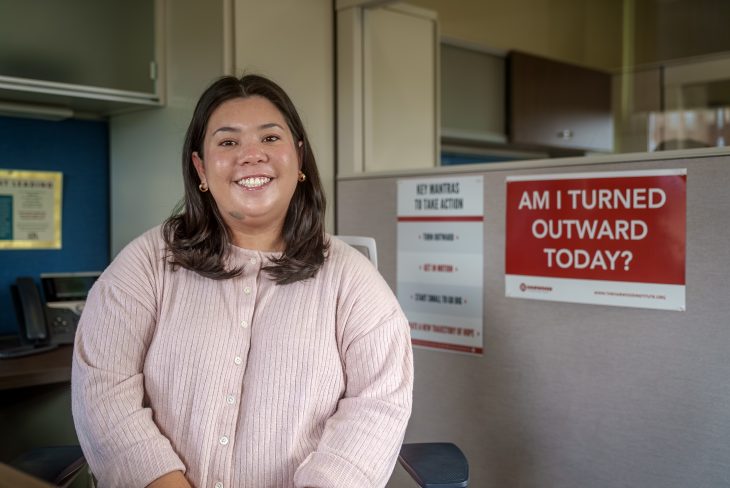 A person sits smiling in an office cubicle, wearing a light pink sweater, with posters on the wall behind them including one that reads “AM I TURNED OUTWARD TODAY?”