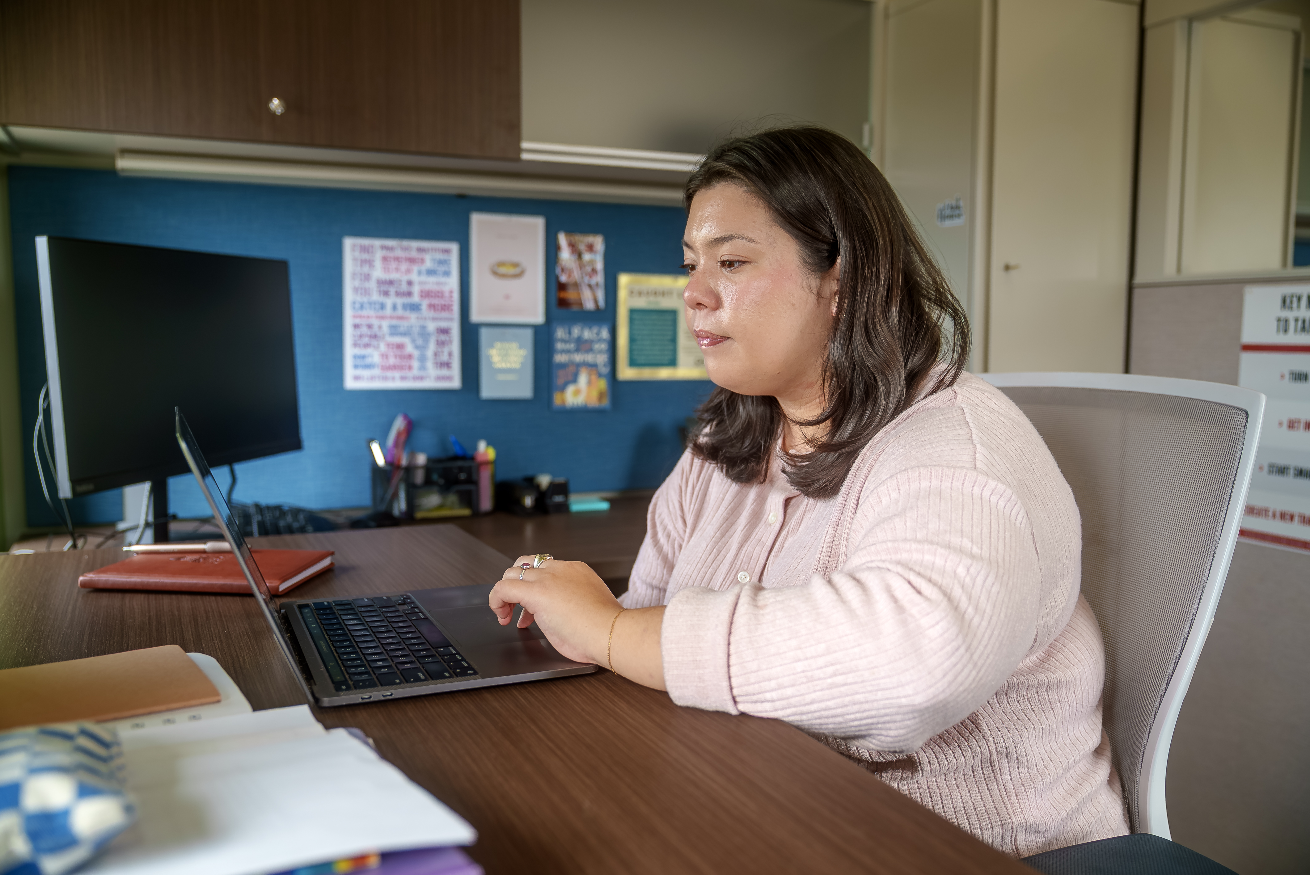 A person sits at a desk in an office, focused on working on a laptop, with papers and notebooks nearby and colorful posters pinned to the wall behind them.