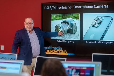 A faculty member gestures toward a screen comparing DSLR and smartphone cameras during a multimedia class.