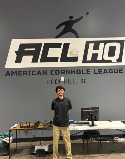 A person stands smiling in front of a wall with the American Cornhole League logo at ACL Headquarters in Rock Hill, South Carolina, with tables and computer equipment behind them.