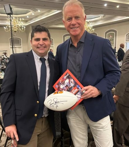 Two people stand together smiling at an indoor event, with one holding a signed football and a booklet as they pose for a photo in a banquet hall.