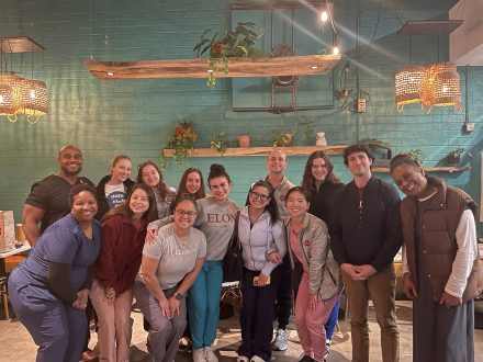 A smiling group of people, some in medical scrubs and others in casual clothing, pose together indoors against a teal brick wall with plants and warm hanging lights.