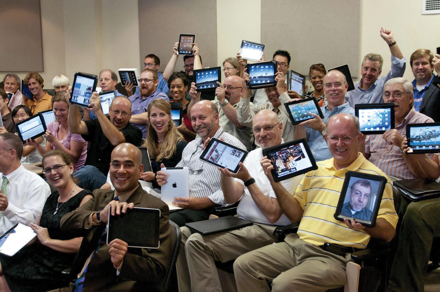 Faculty and staff hold up tablet computers during a workshop, smiling and celebrating new technology.