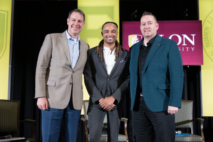 Three men in business attire pose for a photo on stage in front of Elon University banners at the Winter Symposium.