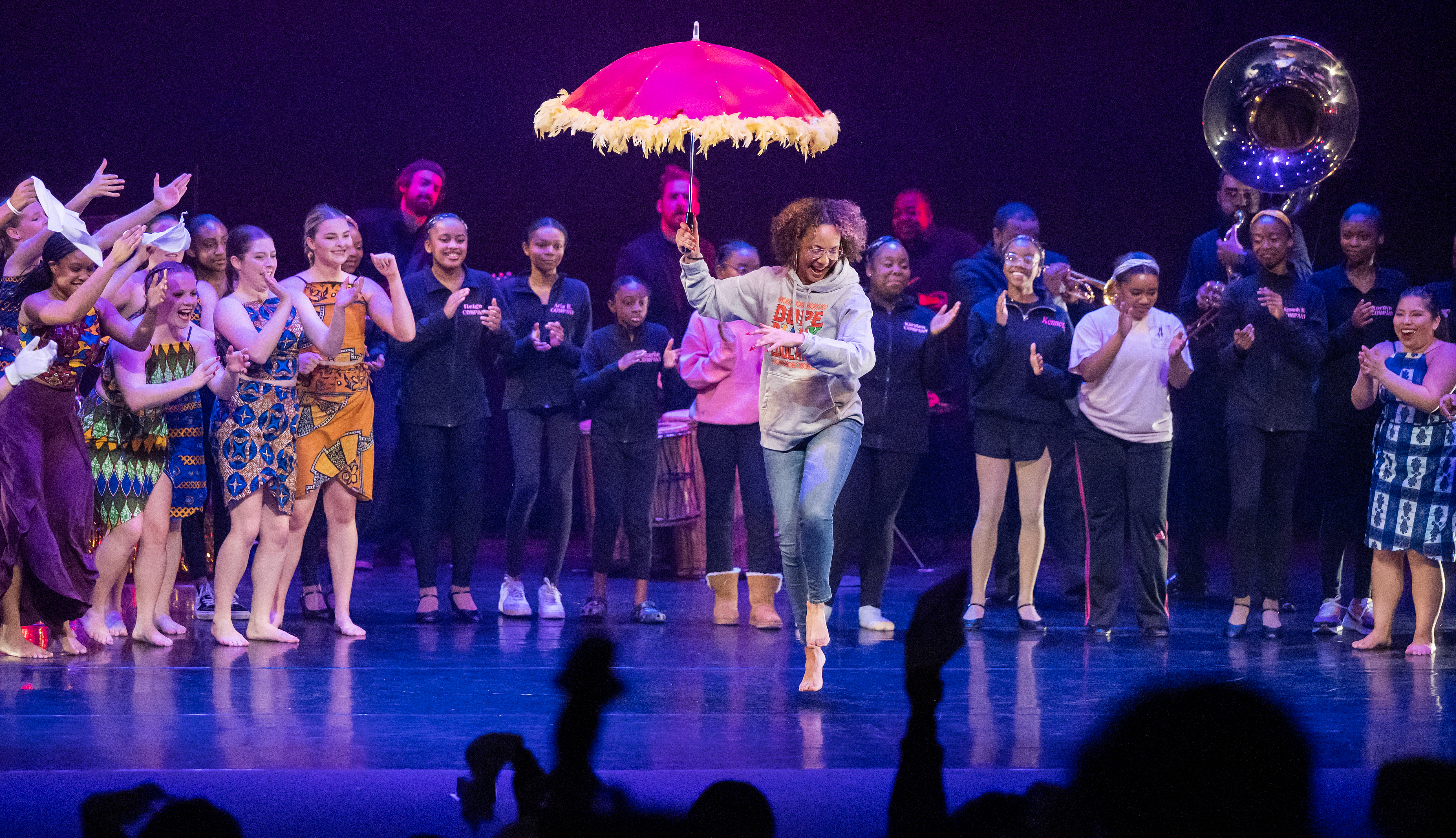 A student dances barefoot across the stage holding a pink umbrella as performers clap and cheer behind her during a Black History Month dance performance.