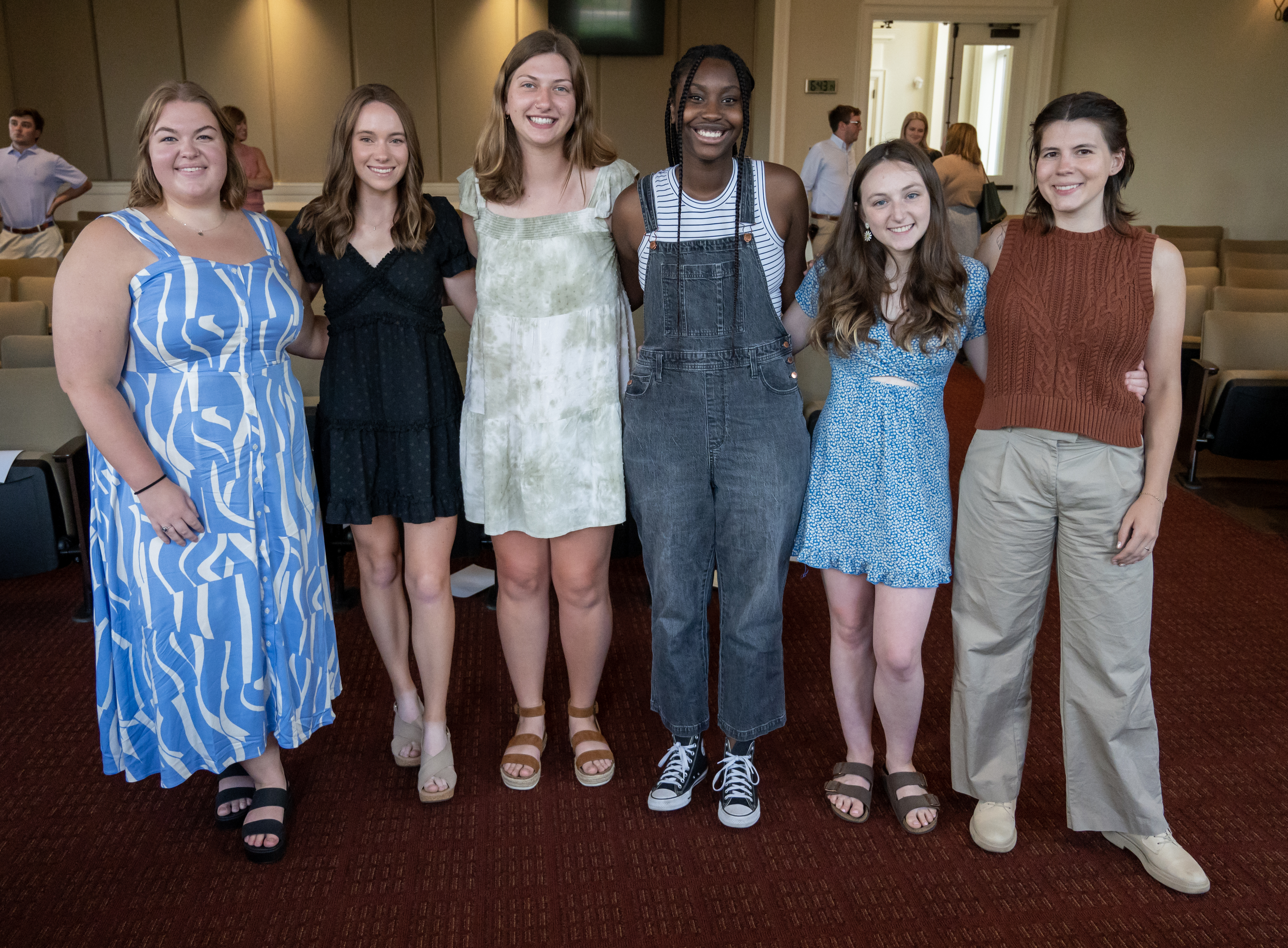 Seven students stand side by side in a campus event room, smiling at the camera. They are dressed in semi-casual attire and pose together in front of rows of chairs, with other attendees visible in the background.