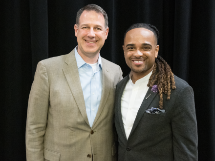 Two men stand side by side, smiling for a photo against a black backdrop.