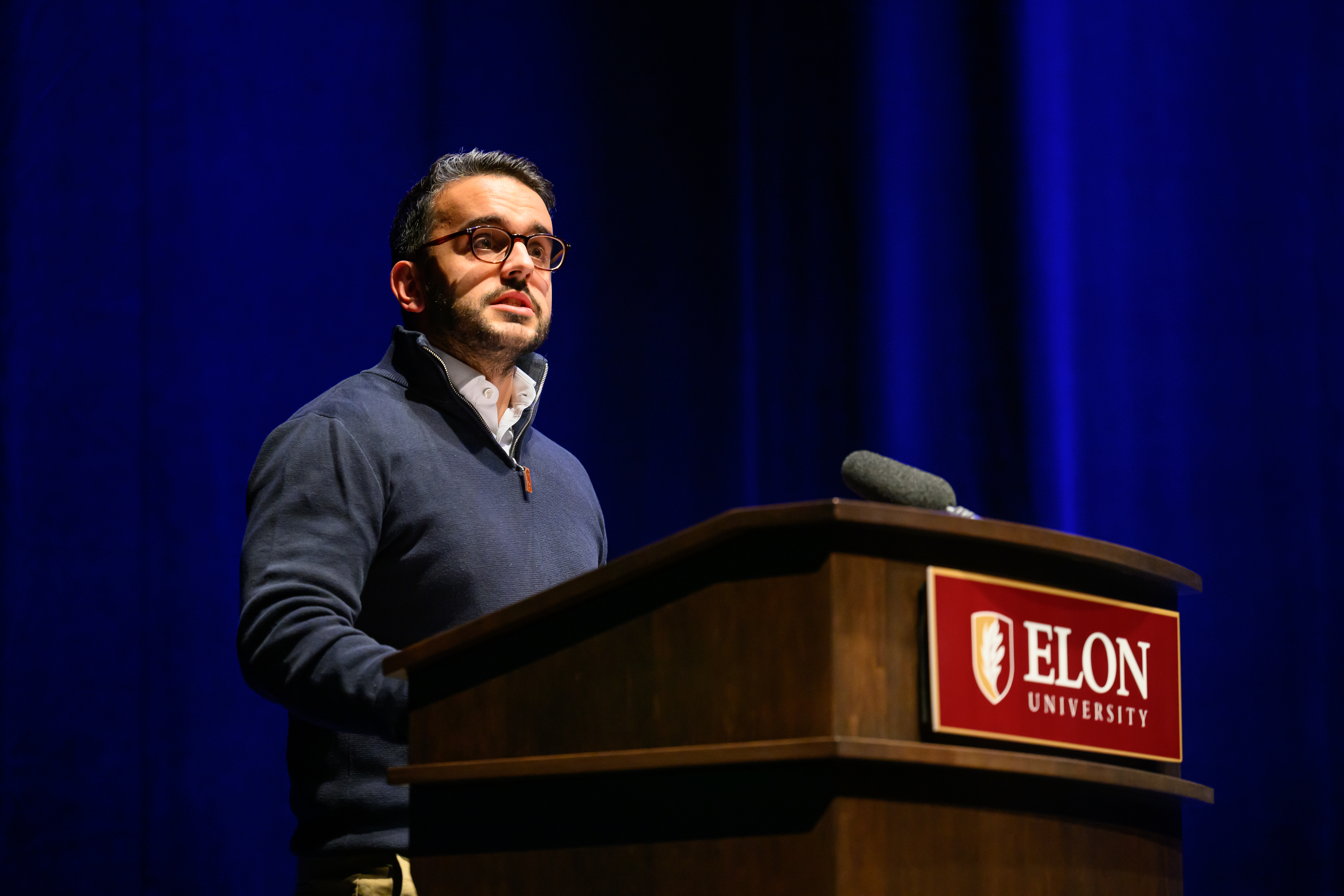 A man in grey sweater speaks behind an Elon University podium