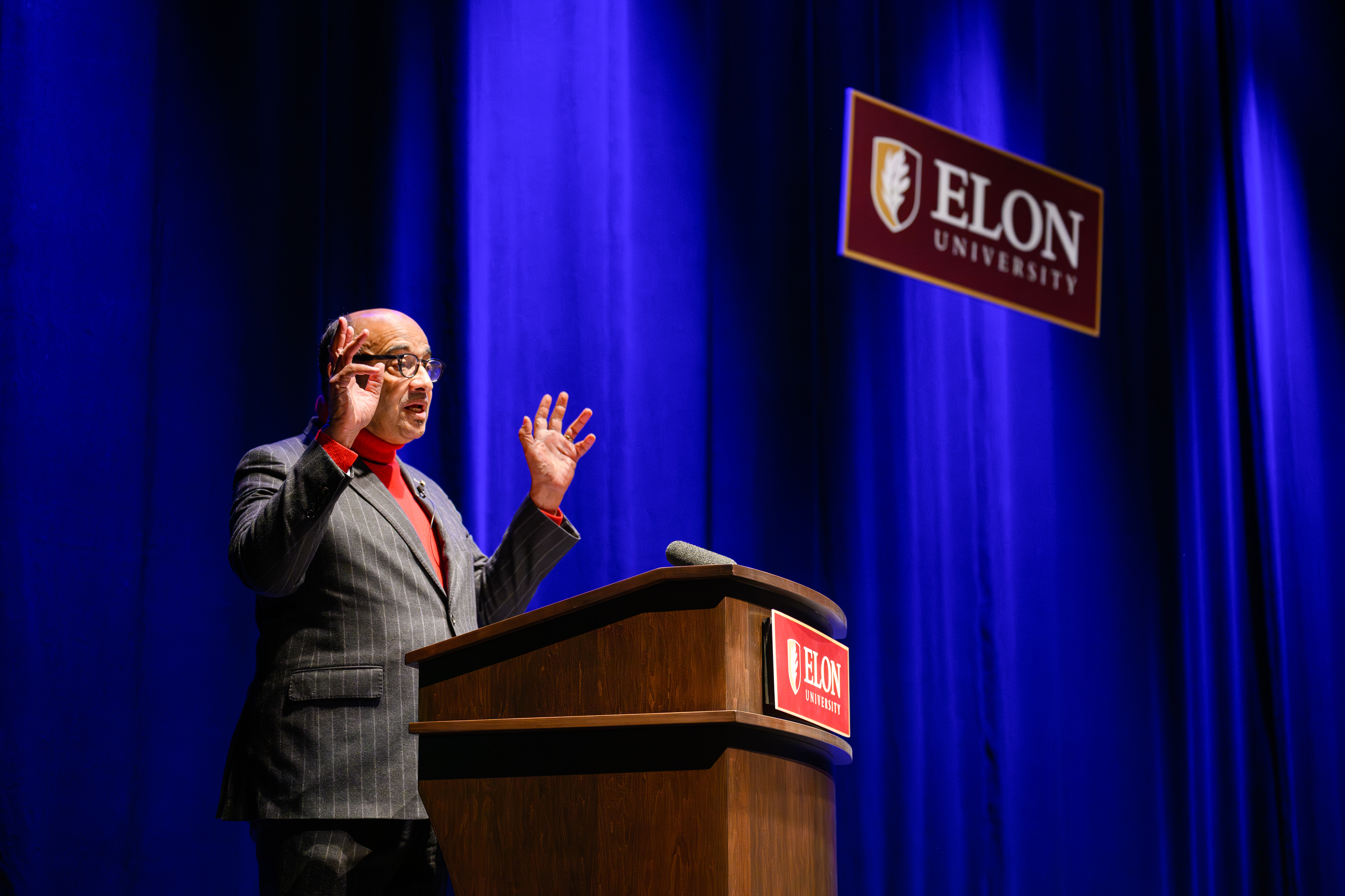 A man with glasses, red turtleneck and grey blazer speaks in front of an Elon University podium